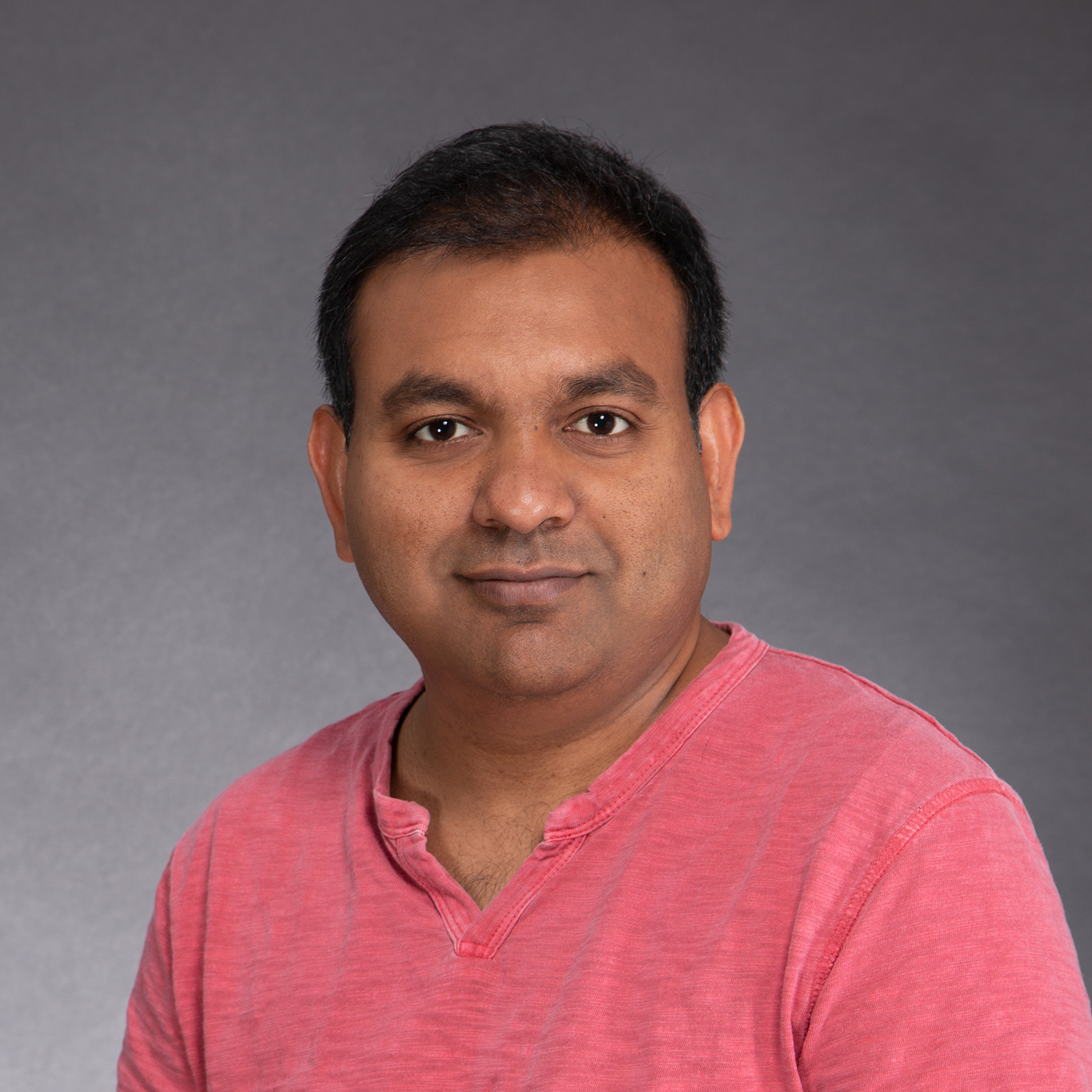 A man with short black hair is facing the camera, wearing a pink V-neck T-shirt. The background is a plain, muted gray color.