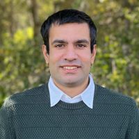 A person wearing a green sweater over a white collared shirt, standing outdoors with trees in the background.